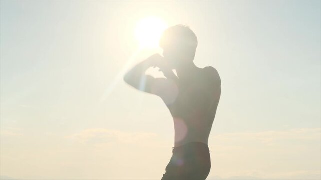 Young muscular fighter trains shirtless alone at sunset, practicing an elbow strike during shadow boxing. MMA, self defense, martial arts training, personal safety and confidence.