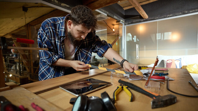 Focused craftsman measuring and marking wooden board among tools at garage workbench. Concept of woodworking preparation, cut list planning, building material estimation.