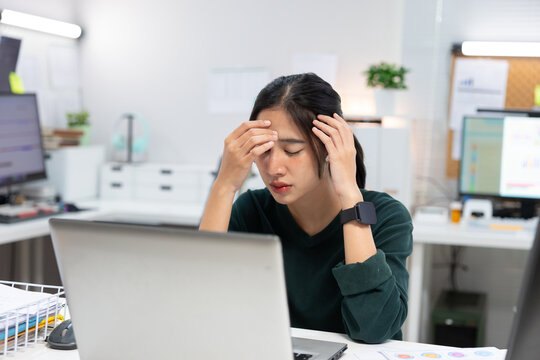 stressed young Asian businesswoman sits at her office desk with her hands on her forehead while looking at a laptop screen.image represents workplace stress, mental fatigue, burnout, deadline pressure