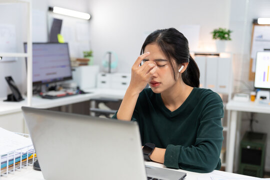 stressed young Asian businesswoman sits at her office desk with her hands on her forehead while looking at a laptop screen.image represents workplace stress, mental fatigue, burnout, deadline pressure