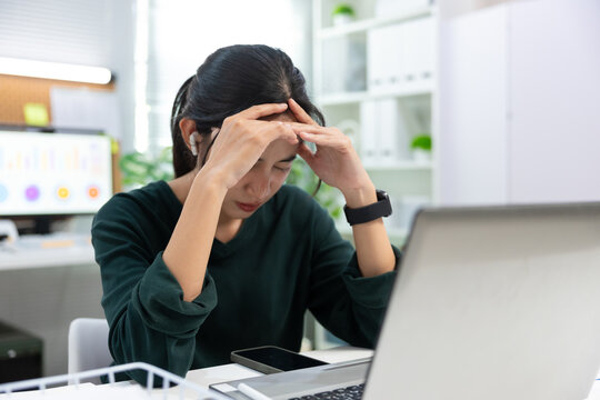 stressed young Asian businesswoman sits at her office desk with her hands on her forehead while looking at a laptop screen.image represents workplace stress, mental fatigue, burnout, deadline pressure