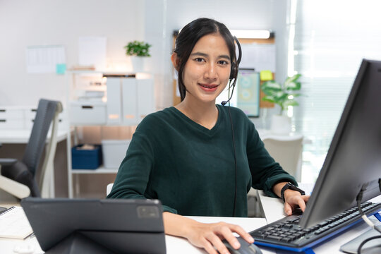 Smiling female customer service representative wearing a headset while working at a desktop computer in a modern office. Confident support agent looking at the camera, providing online customer servic