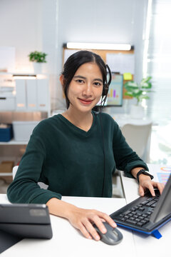 Smiling female customer service representative wearing a headset while working at a desktop computer in a modern office. Confident support agent looking at the camera, providing online customer servic