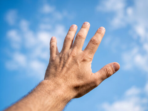 Hand reaching up high towards blue sky with clouds in sun