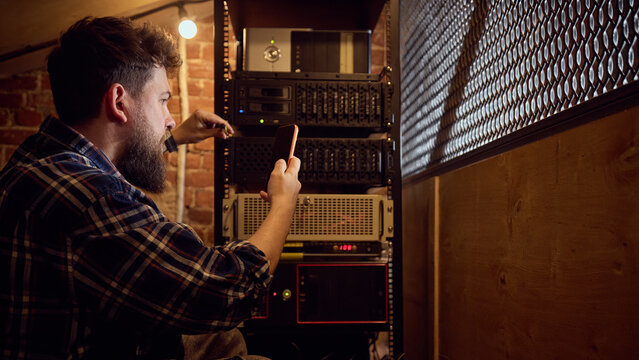 Male technician verifying server rack status with mobile device in technical room. Concept of remote system administration, network performance control, digital infrastructure management.