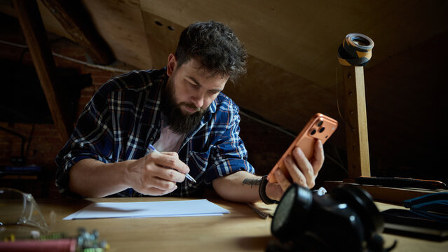 Focused bearded man comparing tools online on smartphone while planning DIY project. Concept of hardware ecommerce, mobile tool shopping, home renovation planning.