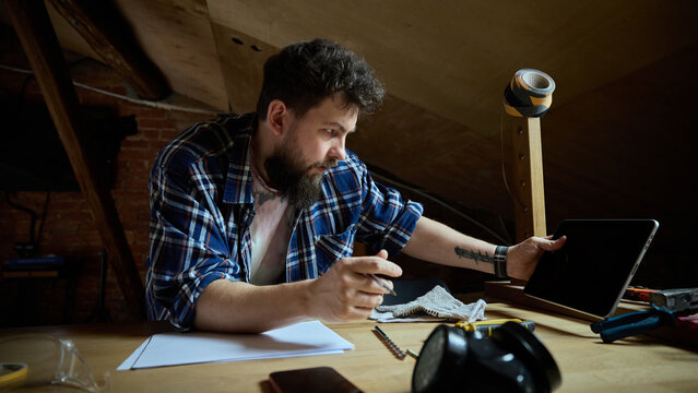 Young craftsman searching workshop supplies on smartphone at wooden workbench. Concept of tool retailer app, online product comparison, repair budget planning.
