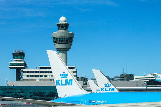 KLM Boeing 737 aircraft tails at Amsterdam Schiphol Airport framed by the airport's air traffic control towers and terminal buildings beneath a bright blue sky.