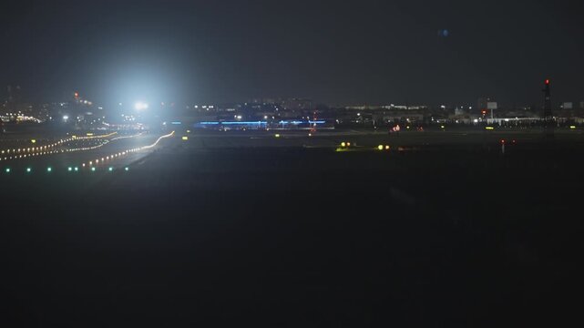 Airport runway illuminated by colorful navigation lights with a windsock and distant terminal buildings at night, airplane landing
