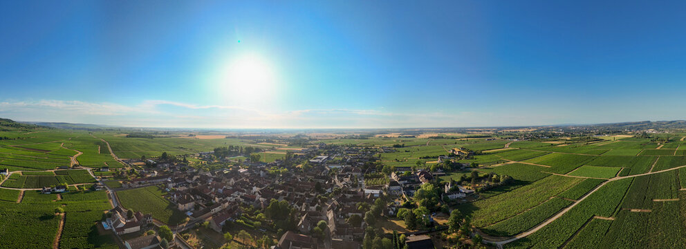 Aerial panorama of a village surrounded by Burgundy vineyards in France, with rows of grapevines stretching across the landscape beneath a bright afternoon sun.