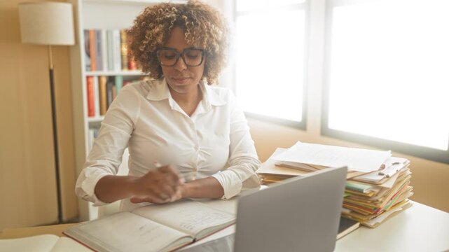 Woman writes in notebook at desk with laptop and tall stack of folders beside her, wearing glasses and white shirt inside a building study; concentration productivity.