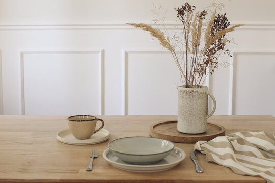 Minimalist Wooden Table Setting with Ceramic Dinnerware, Dried Flowers Grass in Jug Vase. Neutral Scandinavian Dining Room Interior. Cup of coffee, tea. Plate with fork and knife, tea towel.
