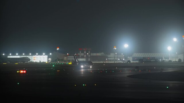 Narrow body aircraft taxiing on night apron, tilt shift lens effect creating selective focus, colorful ramp light and bokeh, terminal and hangar illuminated in background