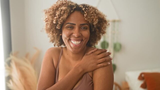 Woman smiling with hand on shoulder, bare shoulder and collarbone visible, wearing a bra in a bright bedroom with plants and pillows; self care joy.