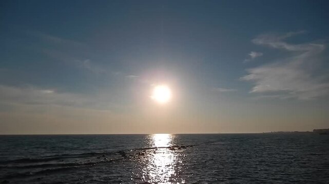 Calm water at sea, moving reflections of sun on small waves at coast with stones and blue sky with clouds on horizon and pathelione. Topics: natural background, travel, sea ​​view, sea horizon