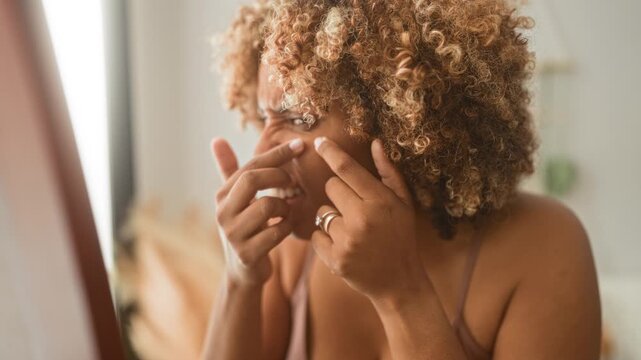 Woman pinches nose blemish with fingers while inspecting skin in studio mirror; self care vulnerability.