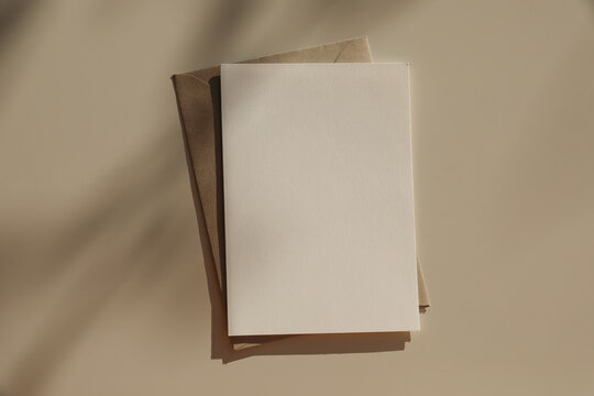 Summer neutral wedding stationery. Closeup of blank greeting card, craft envelope mockup in sunlight. Invitation, template. Beige Table background, soft shadows. Elegant composition. Flatlay, top view