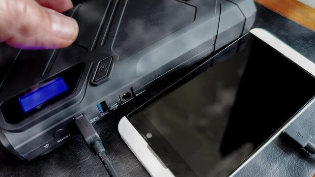 Closeup of a man&rsquo;s hand pressing the button to activate a portable power bank to charge the battery of a mobile phone next to it, connected by USB with a flashing charging light.