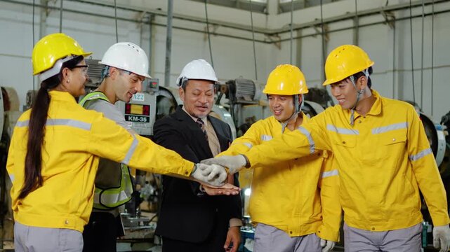 Diverse factory workers and manager in hard hats stack hands together in a huddle, raising them in a cheer and clapping to celebrate success and teamwork inside an industrial manufacturing plant.