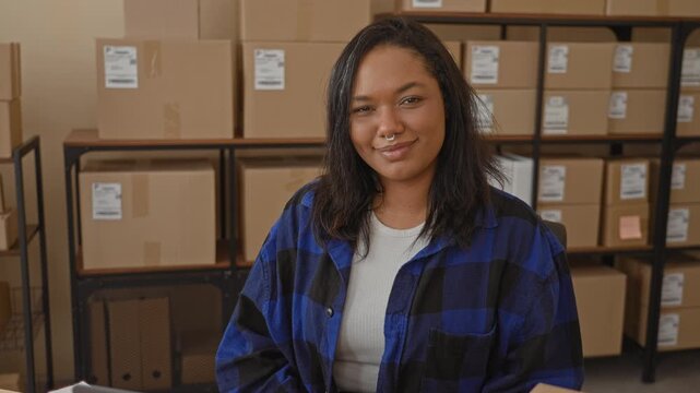 Young woman smiling among cardboard boxes in warehouse storage at a small business shipment desk; entrepreneurial pride.