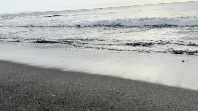 Ocean waves washing onto dark sand beach under overcast sky.