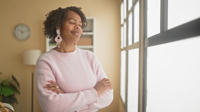 Young african american woman in pink sweater with arms crossed looking out large window in studio, wearing statement earrings and smiling; serenity.