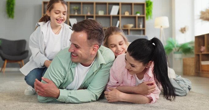 Happy family parents and children playing and tickling. Mom and dad lie on carpet as two daughters share laughter in a cozy living room with light. Concept of family bonding and happiness.