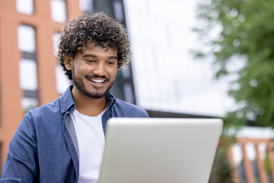 Smiling young man with curly hair focusing on his laptop screen while working or studying outdoors in a modern urban environment, embodying digital nomadism and remote education