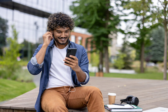 Smiling young man with curly hair sitting outdoors on a university campus and putting on earphones while looking at his mobile phone, creating a moment of relaxation and digital connection