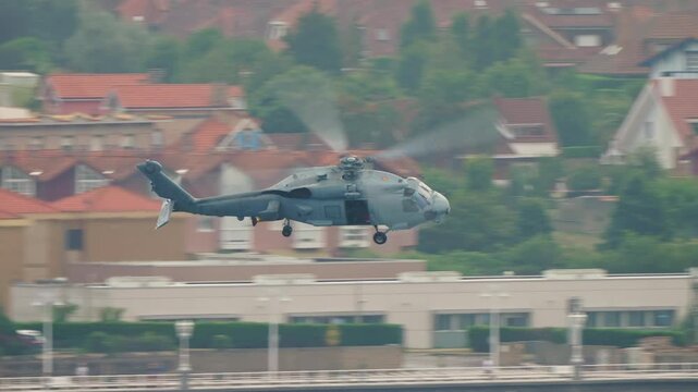 Gray military naval helicopter flies at speed past a large urban building facade with rotor blades blurred, colorful coastal town architecture behind