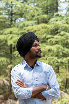 Vertical portrait of a young Sikh man wearing a black turban and a blue shirt, standing with arms crossed while looking away against a blurred background of lush green trees in Himachal Pradesh.