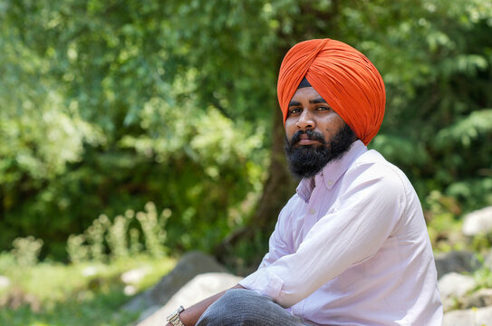 Closeup portrait of a young Sikh man wearing an orange turban, looking directly at the camera while sitting against a blurred background of lush green trees in Himachal Pradesh, with empty copy space.