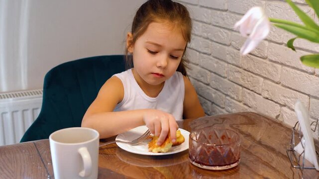 A little Caucasian girl eagerly eats cottage cheese pancakes with berry jam. She enjoys eating foods made with eggs and cottage cheese. Proper nutrition, baby food. Appetite in childhood.