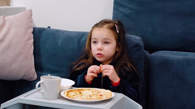 Nutrition for preschool children. Unhealthy food for children. A girl eats pancakes with gusto while sitting on couch at home and watching TV. Unhealthy food for children. Flour pancakes and berry jam
