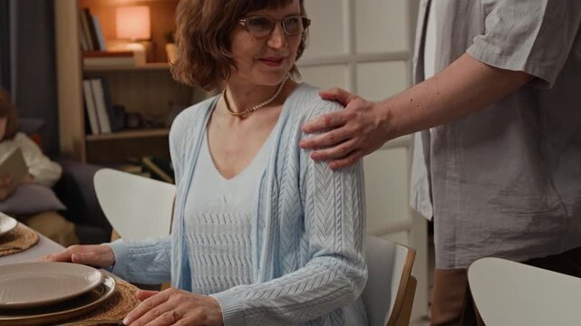 Senior Caucasian woman sitting at dinner table with young adult man assisting with chair. Perfect for family holiday gatherings or domestic lifestyle commercials