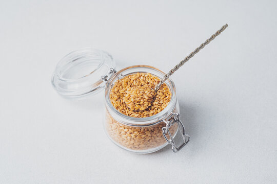 Small glass jar filled with golden flax seeds with a metal spoon inside on a light background