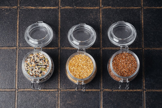 Three clear jars hold mixed seeds, yellow seeds, and brown flax seeds on a dark tiled background