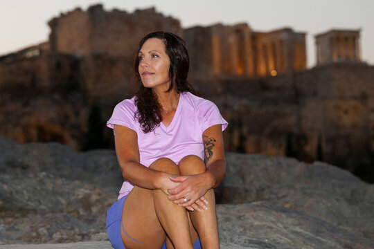 Woman posing on Areopagus Hill with the illuminated Acropolis in Athens, Greece, visible in the background at twilight.