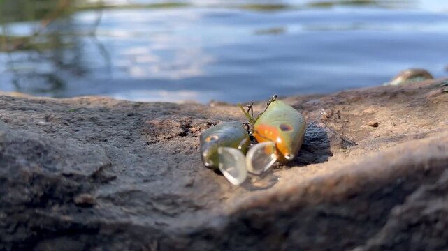 Two hard-body fishing lures with treble hooks rest on a rock beside a fast flowing river in bright late summer light. Close-up with a soft bokeh background