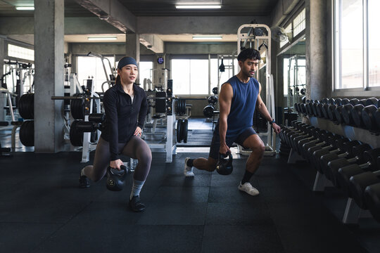 Asian man and woman lifting kettlebell weight inside fitness gym, athletic friends squatting exercise training session strength building workout health lifestyle
