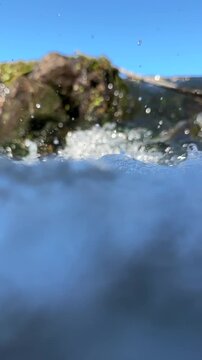 Camera plunges beneath the surface of a fast flowing mountain river, revealing air bubbles, submerged plants and stones in clear sunlit water. Vertical format