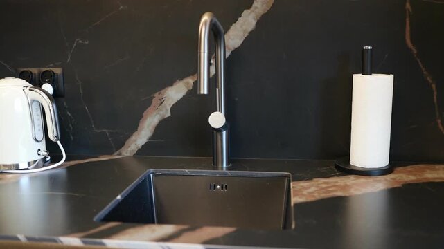 Close up view of a modern chrome water faucet tap over a dark sink in a stylish contemporary kitchen. A white electric kettle and a paper towel holder stand on the dark marble countertop, capturing