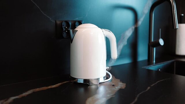 Close up view of a modern white electric kettle with a glowing blue LED indicator light switch, boiling water on a dark marble kitchen countertop. Next to it stands a sleek black faucet tap, capturing