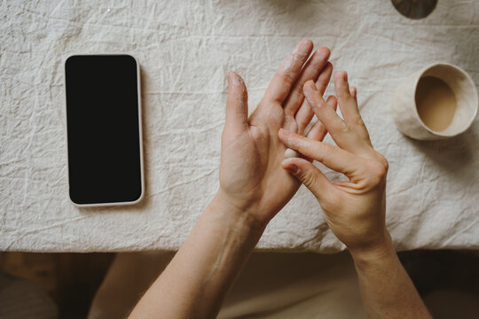 Female hands shaping clay with pottery tools near smartphone.