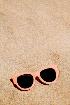 a close-up of pale pink sunglasses on the sand of a beach