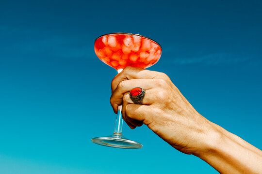 a man raises a glass of red cocktail and ice against a blue sky