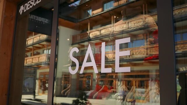 Close up view of a large white 'SALE' sign text sticker applied on the glass window of a modern fashion clothing store. The interior of the retail boutique with clothes on hangers is softly visible