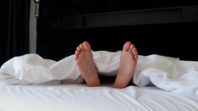 Close up view of bare male feet peaking out from under a soft white blanket or duvet on a bed. In the blurred background, a young man is seen lying down and resting, capturing a comfortable sleep