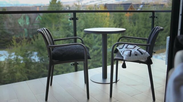 Close up view of an empty outdoor seating area on a hotel balcony or terrace. The patio features a small round table and two comfortable wicker armchairs with cushions, overlooking beautiful alpine