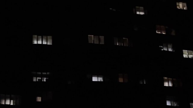 Night view on apartment building with illuminated windows. Warm lights glow from different flats, creating a quiet urban atmosphere and a sense of life behind the facades in the darkness. Slow motion
